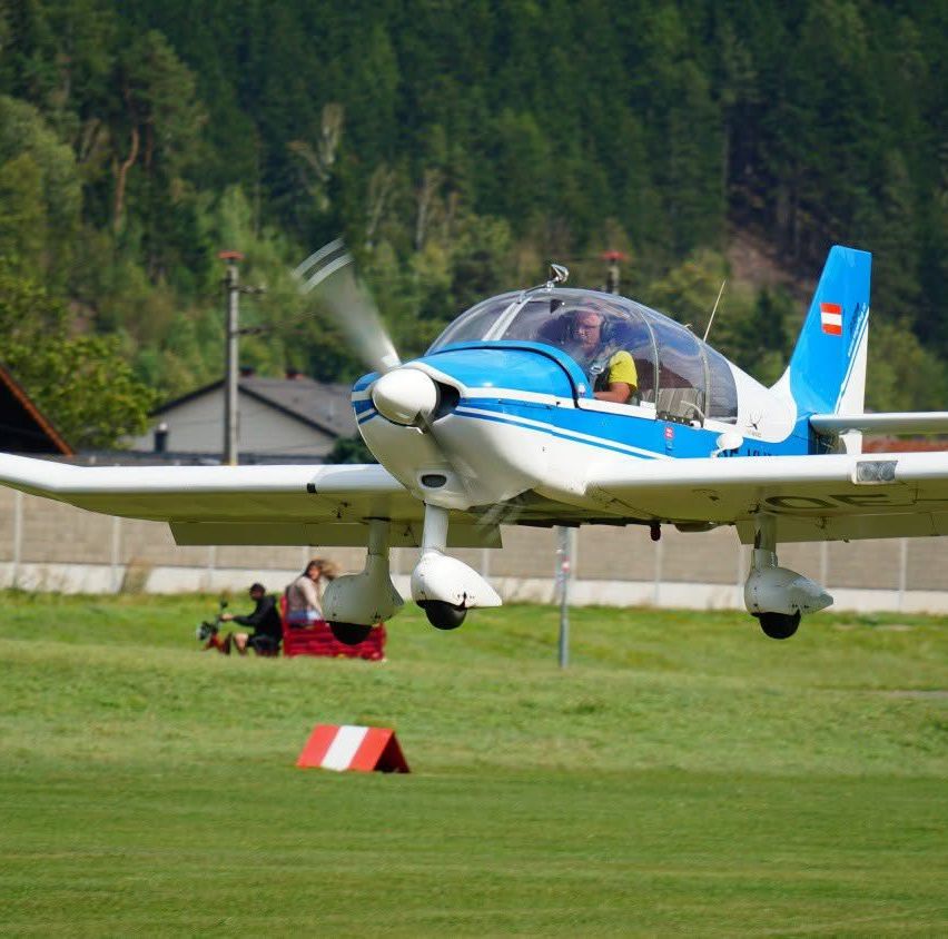 Ein blaues und weißes Flugzeug startet von einem Grasfeld mit einer Person im Inneren. Im Hintergrund befinden sich Bäume und ein Gebäude.