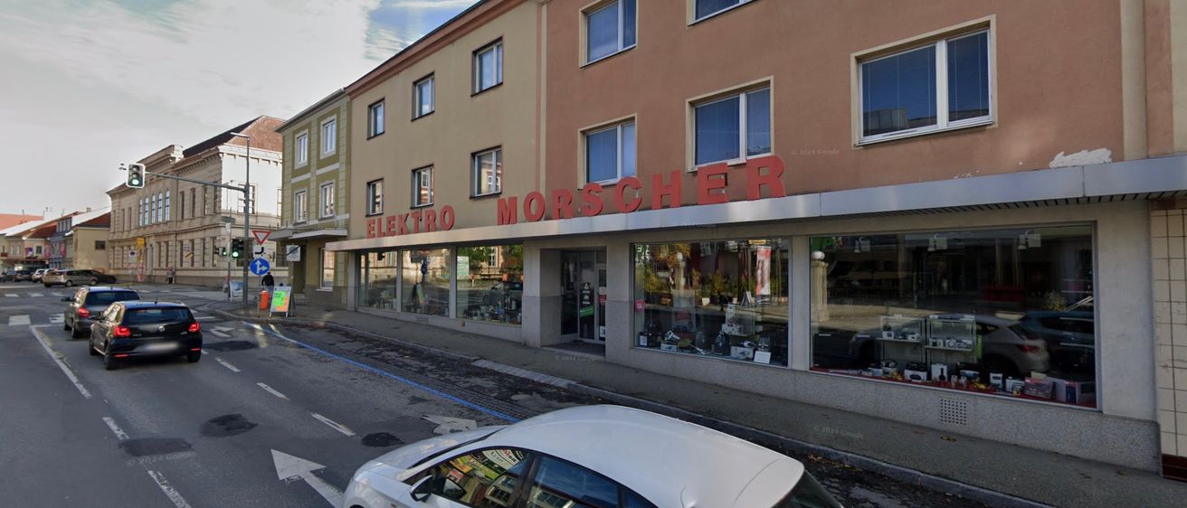 Ein weißer Wagen ist am Straßenrand vor dem Elektro Morscher Geschäft geparkt, das ein rotes Schild und große Glasfenster hat.