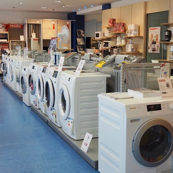 Ein Ausstellungsraum zeigt eine Reihe weißer Waschmaschinen und Trockner, mit verschiedenen Haushaltsgeräten und Werbematerialien rundherum.