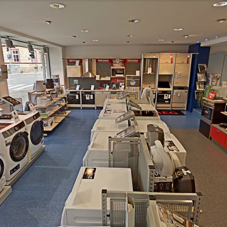 Ein Geräteladen mit Reihen von Waschmaschinen und Öfen zur Ausstellung. Der Boden ist blau und die Decke hat Einbauleuchten.