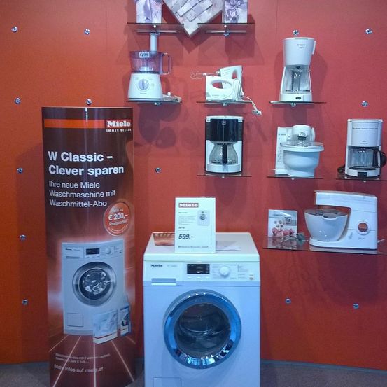 An einer roten Wand ist eine Ausstellung weißer Haushaltsgeräte zu sehen. Ein großer zylindrischer Poster für Miele steht neben einer Waschmaschine. Regale halten verschiedene Geräte, darunter einen Mixer, eine Kaffeemaschine und einen Mixer.