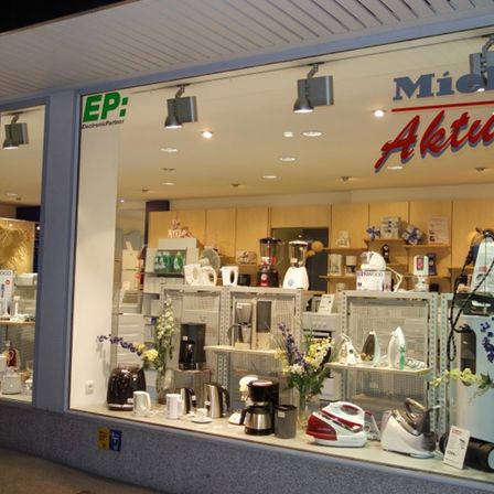Ein Schaufenster zeigt verschiedene Haushaltsgeräte wie Kaffeemaschinen, Wasserkocher und Bügeleisen, die auf Regalen angeordnet sind. Der Ladenname 'Miele Aktu' ist prominent im Glas angebracht.