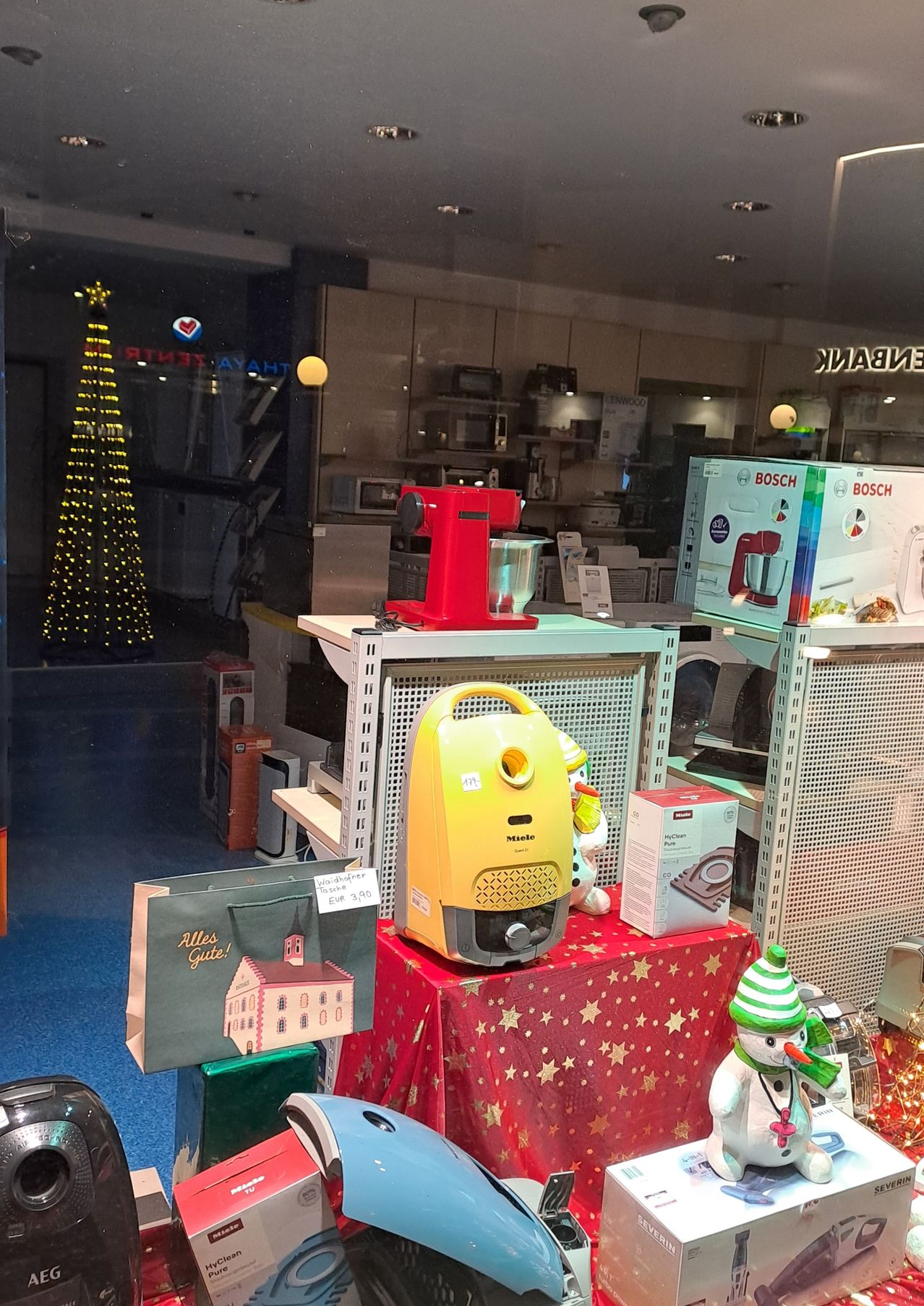 Ein Schaufenster zeigt verschiedene Haushaltsgeräte, darunter einen roten Mixer, einen gelben Staubsauger und mehrere Kisten. Ein Weihnachtsbaum und ein Schneemann sind ebenfalls zu sehen. Das Fenster hat ein Weihnachtsthema mit Dekorationen und einem Beutel mit 'Alles Gute!' geschrieben.