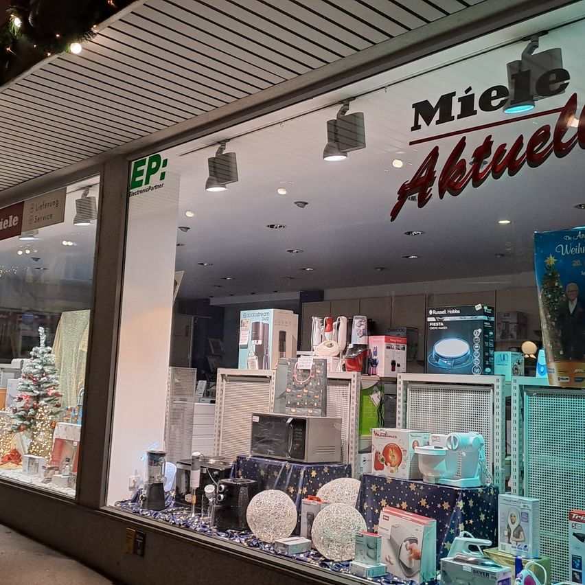 Ein Schaufenster zeigt Weihnachtsartikel und Haushaltsgeräte, mit einem Schild, das 'Miele' und 'Aktuell' anzeigt. Im Inneren sind verschiedene Produkte auf Regalen und ein Weihnachtsbaum in der Ecke zu sehen.