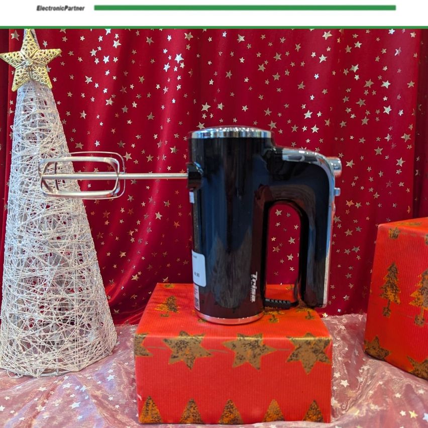 Ein schwarzer Mixer steht auf einem roten Geschenkkarton mit Weihnachtsdekoration im Hintergrund. Der Karton hat ein Sternenmuster. Links steht ein weißer Weihnachtsbaum mit Sternen.