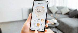 Eine Person hält ein Smartphone, das eine Temperaturanzeige von 22 Grad Celsius zeigt. Der Bildschirm des Telefons zeigt verschiedene Symbole, möglicherweise im Zusammenhang mit der Hausautomation.