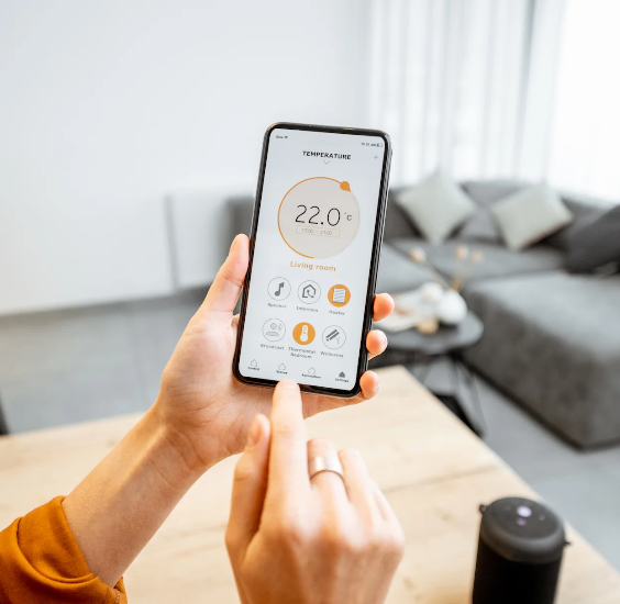 Eine Person hält ein Smartphone, das eine Temperaturanzeige von 22,0 Grad Celsius zeigt. Der Bildschirm zeigt Symbole für ein Wohnzimmer, Lautsprecher, Thermostat und mehr. Der Hintergrund umfasst eine Couch und einen Couchtisch.