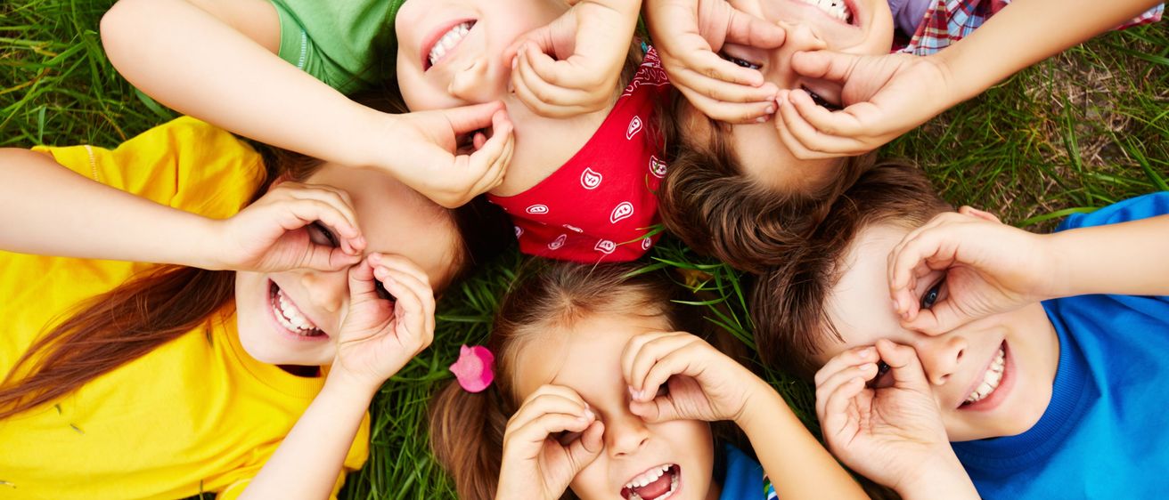 Fünf Kinder liegen auf dem Gras, lächeln und posieren für ein Foto. Sie haben ihre Hände auf den Augen und bilden einen Kreis um ein Mädchen in einem roten Shirt.