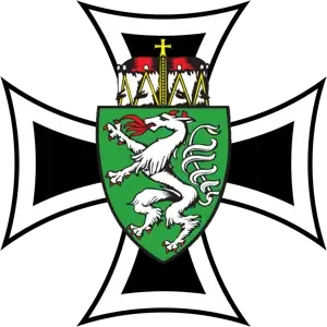 Ein heraldisches Emblem mit einem schwarzen Kreuz und einem weißen Wolfswappen, das einen rotzüngelnden Wolf auf einem grünen Schild mit einem Kreuz auf der Oberseite zeigt, vor schwarzem Hintergrund.