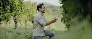 Ein Mann kniet in einem Weinberg, hält eine Ringbox und hat einen freudigen Ausdruck, umgeben von grünen Blättern.