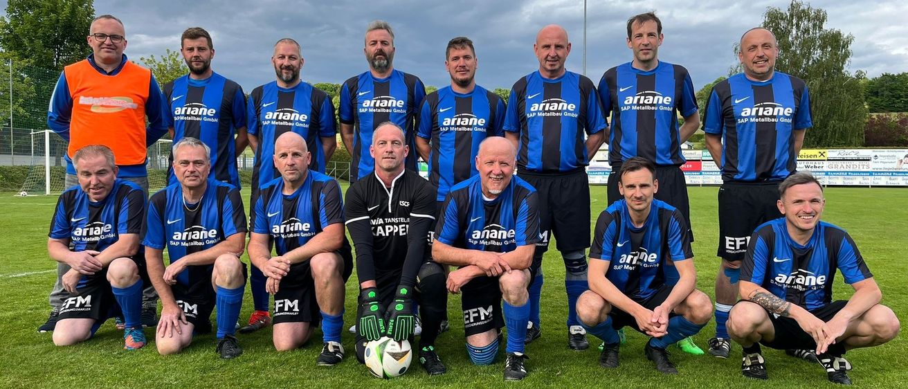 Eine Fußballmannschaft in blauen und schwarzen Trikots mit dem Wort 'ariane' darauf posiert für ein Gruppenfoto auf einem Rasenfeld unter einem bewölkten Himmel.