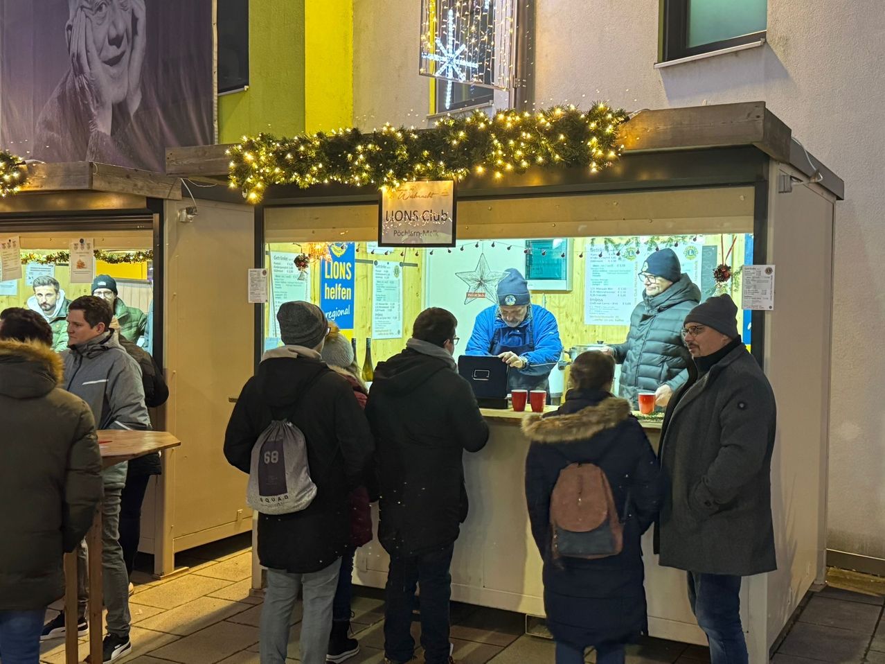 Menschen stehen in einer Schlange vor einem kleinen Kiosk, wahrscheinlich um Essen oder Getränke zu kaufen. Der Kiosk hat eine Weihnachtsgirlande und ein Schild, das Lions Club steht.