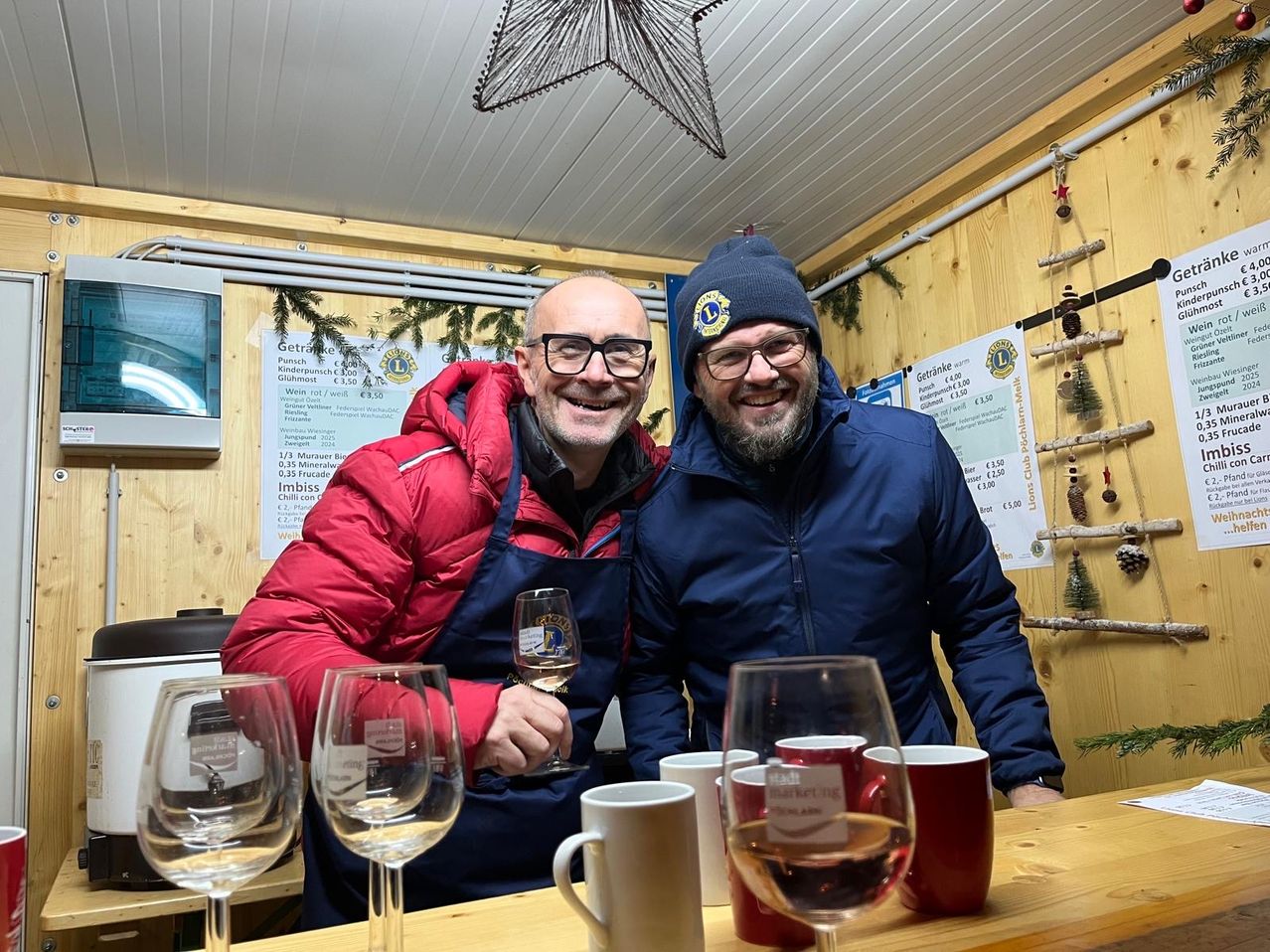 Zwei Männer stehen an einer Bar mit Weingläsern und Tassen. Einer hält ein Glas Wein, der andere lächelt. Hinter ihnen zeigt eine Wand Artikel zum Verkauf an, und Dekorationen hängen.