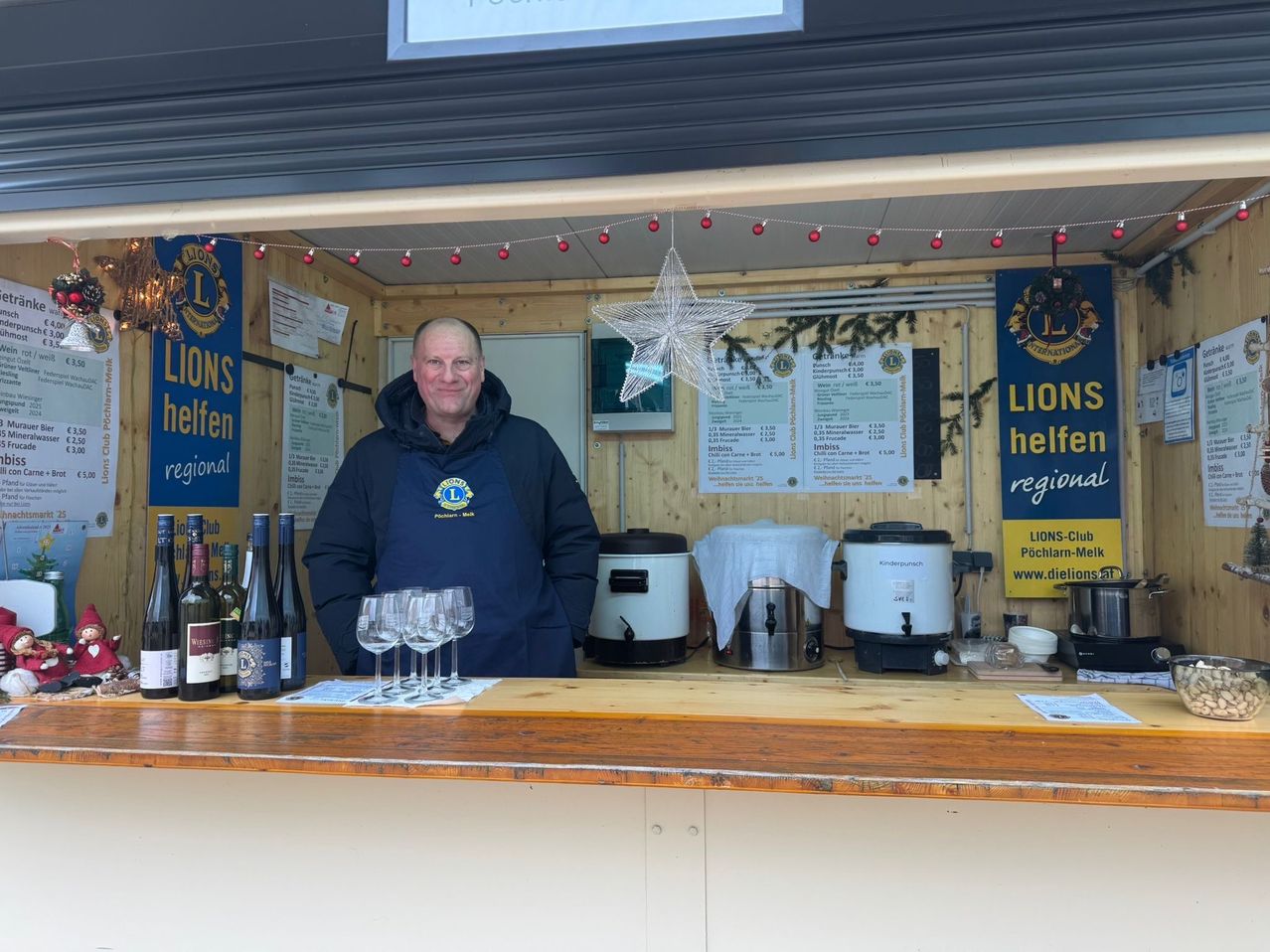 A man stands behind a counter with bottles and wine glasses, next to a coffee machine. A star hangs from the ceiling, and there are signs for Lions helfen regional.