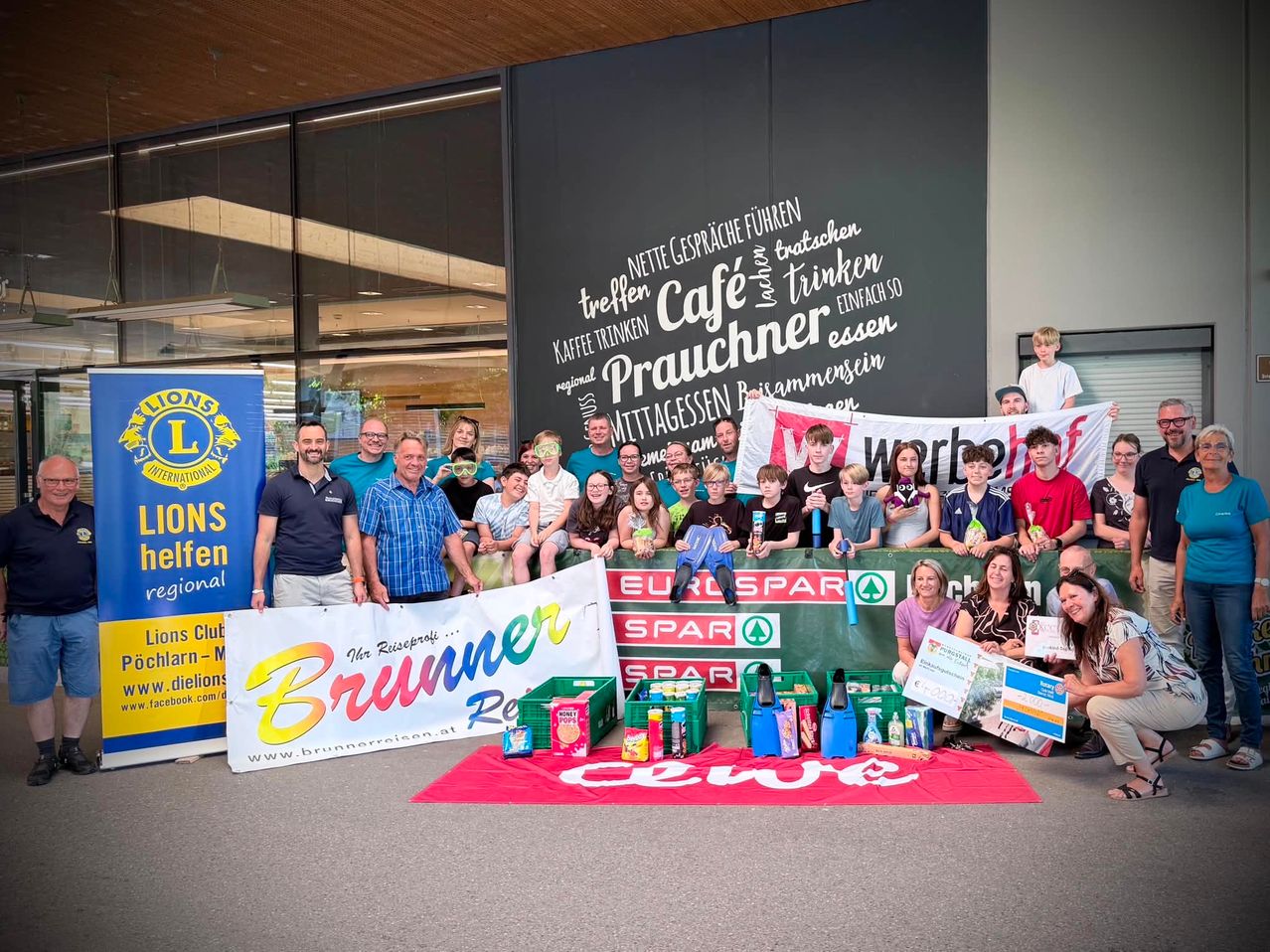 A group of people, possibly volunteers, pose with a banner and various goods in front of a wall. They are smiling and appear to be at a charity event.