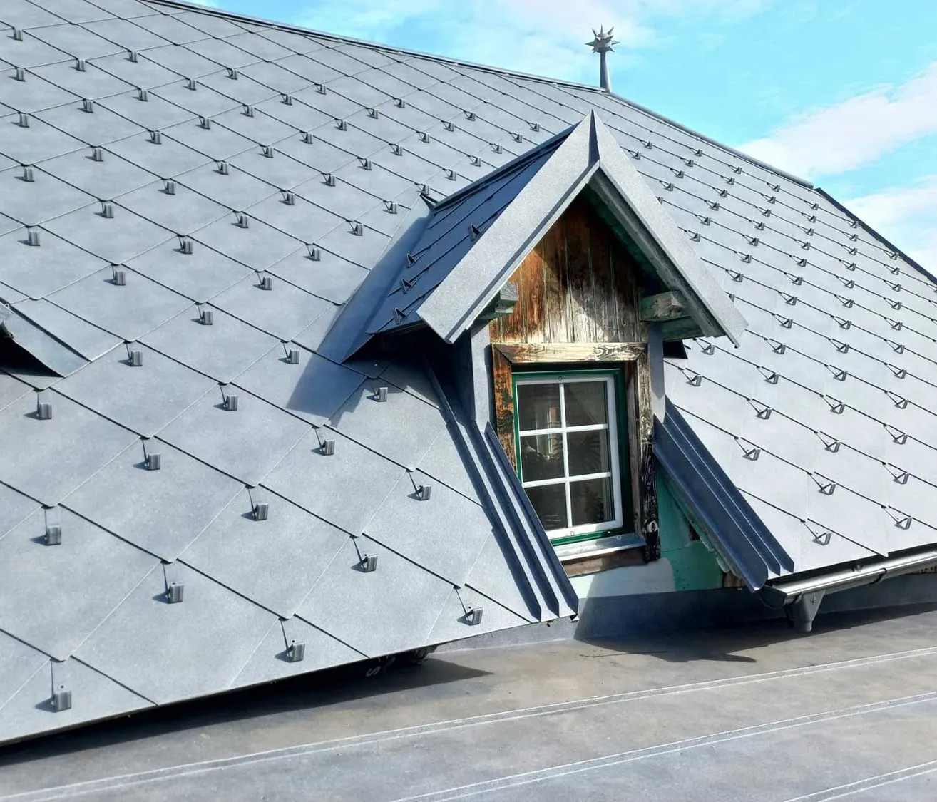 Ein Dach mit einem dreieckigen Giebel, das ein Fenster mit hölzerne Rahmen und Glasscheiben zeigt. Das Dach ist mit grauen Schindeln bedeckt.