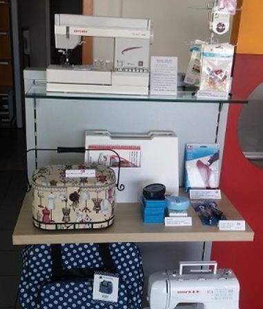 Ein Ausstellungsregal mit verschiedenen Nähmaschinen, einer Punkttasche und anderen Artikeln. Eine weiße Nähmaschine ist prominent ausgestellt, zusammen mit einer gemusterten Tasche und zusätzlichen Zubehörteilen.