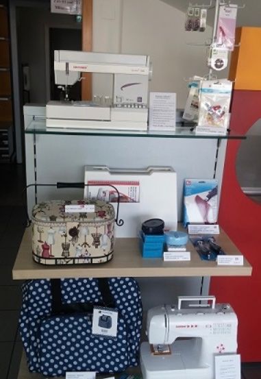 Ein Ausstellungsregal mit verschiedenen Nähmaschinen, einer Punkttasche und anderen Artikeln. Eine weiße Nähmaschine ist prominent ausgestellt, zusammen mit einer gemusterten Tasche und zusätzlichen Zubehörteilen.
