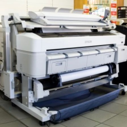 Ein Epson-Drucker ist in einem Raum mit gefliestem Boden neben einem anderen Drucker ausgestellt. Der Epson-Drucker hat einen großen, weißen Körper mit einer blauen Plattform und einem Papierrollen.