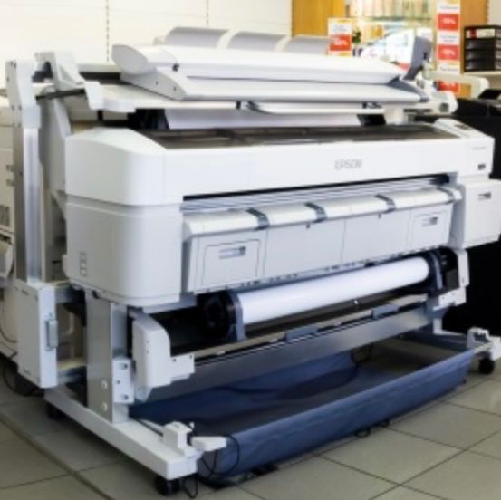 Ein Epson-Drucker ist in einem Raum mit gefliestem Boden neben einem anderen Drucker ausgestellt. Der Epson-Drucker hat einen großen, weißen Körper mit einer blauen Plattform und einem Papierrollen.
