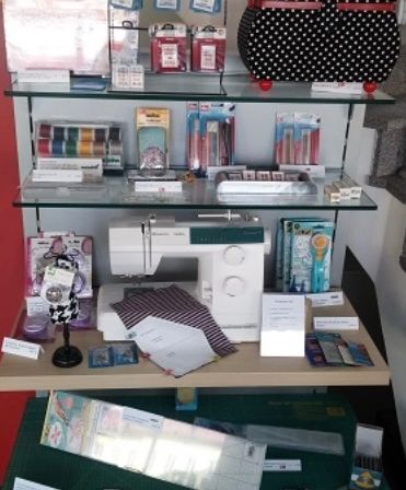 Ein Displaytisch mit Regalen voller Nähzubehör, einschließlich einer Nähmaschine, Stoff und verschiedenen Werkzeugen. Oben steht eine Tasche mit Punkten und unten eine grüne Schneidematte.