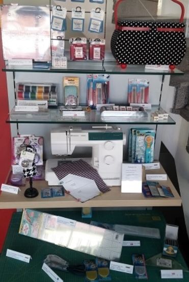 Ein Displaytisch mit Regalen voller Nähzubehör, einschließlich einer Nähmaschine, Stoff und verschiedenen Werkzeugen. Oben steht eine Tasche mit Punkten und unten eine grüne Schneidematte.