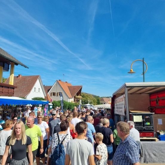 Ein geschäftiger Outdoor-Markt an einem sonnigen Tag. Menschen gehen herum, einige mit Rucksäcken, und es gibt Stände mit ausgestellten Artikeln. Der Himmel ist blau mit einigen Wolken.