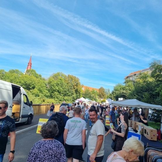 Ein Markt im Freien mit mehreren Ständen unter weißen Zelten. Menschen gehen vorbei und betrachten die Artikel. Ein Lieferwagen ist geparkt. Bäume und Gebäude sind im Hintergrund.