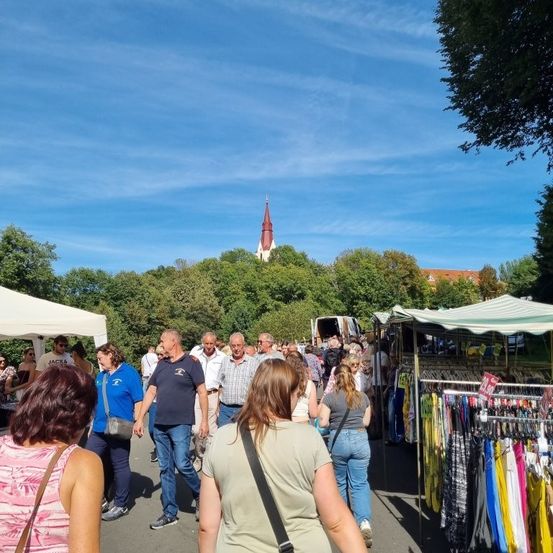 Eine Gruppe von Menschen schlendert durch einen Markt mit Ständen unter Zelten, bunten Kleidern zur Schau gestellt und einer Kirchturm im Hintergrund.