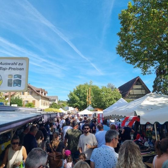 Ein geschäftiger Outdoor-Markt mit zahlreichen Ständen unter weißen Zelten. Menschen gehen herum, einige kaufen ein. Ein Schild auf der linken Seite steht 'Auswahl zu Top-Preisen'. Bäume und Häuser sind im Hintergrund.
