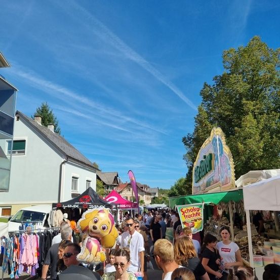 Eine geschäftige Outdoor-Marktszene mit vielen Menschen, Ständen und Bäumen. Ein Hund-förmiger Ballon wird von einer Person gehalten. Ein Haus mit Schornstein ist im Hintergrund.