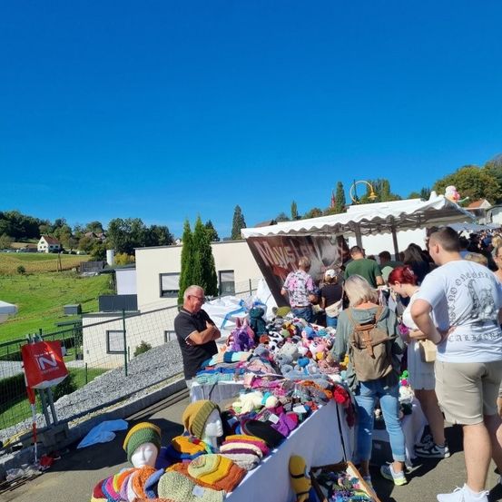 Ein Markt mit einer Vielzahl bunter gestrickter Mützen und Artikeln ist draußen aufgebaut, mit Menschen, die stöbern, und einem Gebäude im Hintergrund.