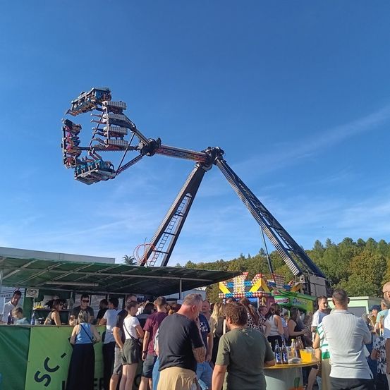 Eine Gruppe von Menschen steht unter einem Vordach in der Nähe einer großen, bunten Schaukel, mit einem klaren blauen Himmel im Hintergrund.