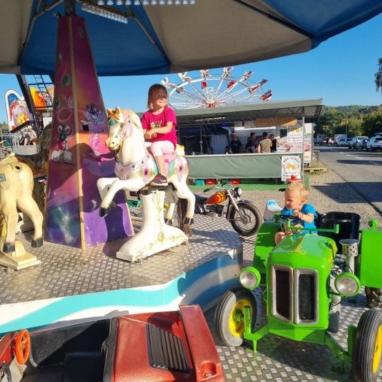 Ein kleines Mädchen sitzt auf einem mechanischen Pferd in einem Vergnügungspark. Ein Baby fährt einen grünen Traktor in der Nähe. Im Hintergrund sind ein Riesenrad und verschiedene Fahrgeschäfte zu sehen.