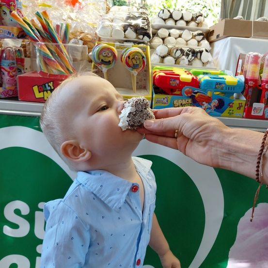 Ein Baby in einem blauen Hemd isst einen mit Schokolade überzogenen Donut, gehalten von einem Erwachsenen mit einer Armband. Dahinter zeigt ein Süßigkeiten-Tresen verschiedene Leckereien.