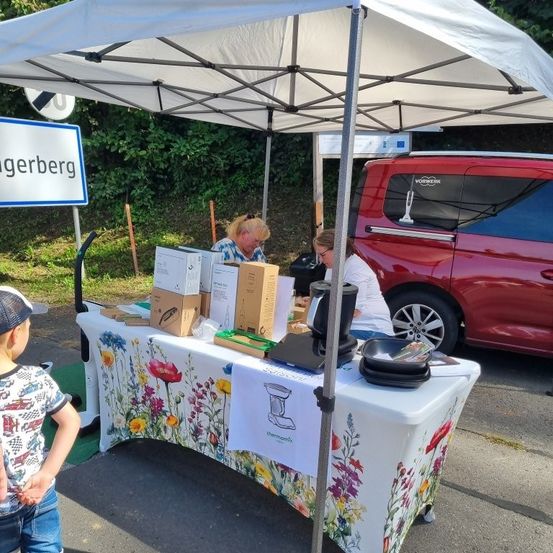 Zwei Frauen arbeiten unter einem Zelt. Sie verkaufen Produkte aus Kisten auf einem Tisch. Ein junger Junge steht in der Nähe. Ein roter Van ist hinter ihnen geparkt.