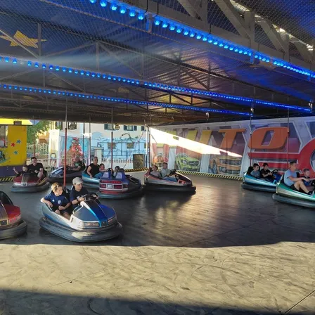 Mehrere Autoscooter fahren unter einer blau beleuchteten Decke in einem Vergnügungspark. Mehrere Personen genießen die Fahrt.