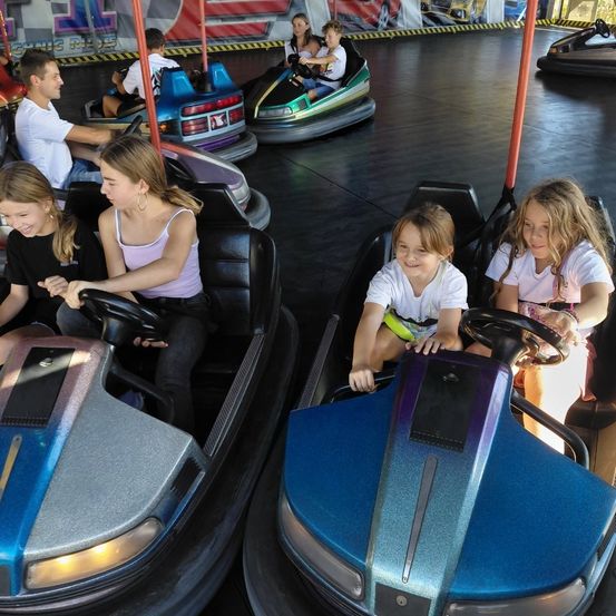 Mehrere Autoscooter sind mit Kindern und Erwachsenen gefüllt. Sie haben Spaß und lächeln. Die Autoscooter sind bunt und auf einem glänzenden Boden.