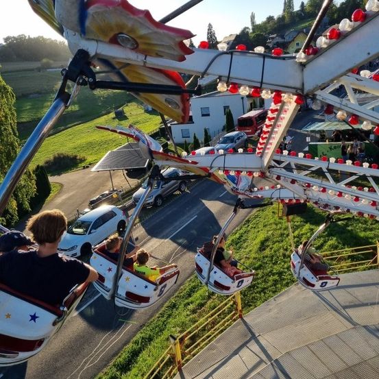 Luftaufnahme einer Karussellfahrt mit vier Sitzen. Die Fahrgäste sitzen auf den Sitzen, während die Fahrt in Bewegung ist. Im Hintergrund sind geparkte Autos, Häuser und eine Straße zu sehen.