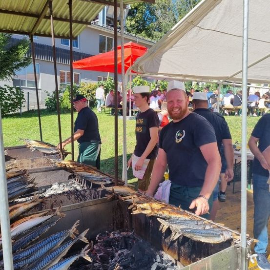 Eine Gruppe von Menschen bei einem Grillfest im Garten, wo sie Fische unter einem Zelt grillen. Zwei Männer tragen Kochmützen, und andere sitzen an Tischen im Hintergrund.