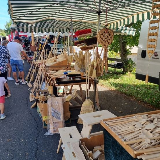 Ein Outdoor-Markt mit einem grün-weißen Baldachin, der verschiedene handgefertigte Holzbesen, Utensilien und andere Gegenstände auf Tischen und Kisten darunter ausstellt. Menschen schauen sich die Artikel an.