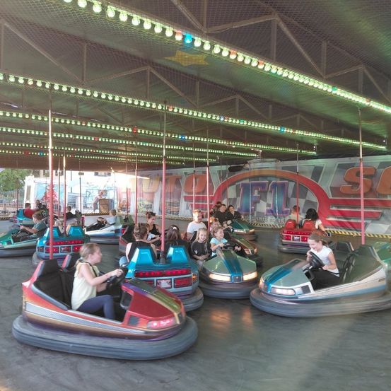 Eine Gruppe von Menschen fährt im Indoor-Vergnügungspark auf Autoscootern. Bunte Lichter beleuchten die Decke und es gibt ein großes Schild mit den Buchstaben "SF".