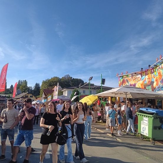 Eine Gruppe von Menschen geht an Essensständen vorbei unter einem klaren blauen Himmel. Rote Fahnen und ein gelber Regenschirm sind sichtbar. Die Stände haben bunte Poster. Leute stehen in einer Schlange an einer Mülltonne.