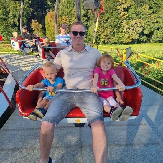 Ein Mann sitzt auf einer roten Fahrt mit zwei Kindern, einem Jungen und einem Mädchen, trägt eine Sonnenbrille und lächelt. Andere Fahrgäste sitzen hinter ihnen. Die Fahrt ist von einem gelben Zaun umgeben, mit einem Grasfeld und Bäumen im Hintergrund.