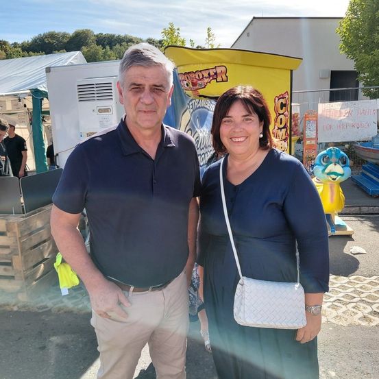Ein Mann und eine Frau stehen lächelnd vor einer Karnevalsattraktion. Hinter ihnen befinden sich Holzkisten und ein weißes Zelt. Der Mann trägt ein dunkles Poloshirt und Khakihosen, die Frau ein marineblaues Kleid und trägt eine weiße Tasche. Im Hintergrund ist ein blau-gelber aufblasbarer Vogel zu sehen.