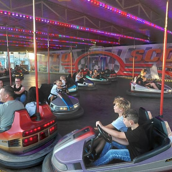 Mehrere Bumper Cars mit bunten Lichtern an der Decke. Mehrere Menschen fahren sie, darunter zwei Jungen am Steuer. Die Wände haben große Logos.