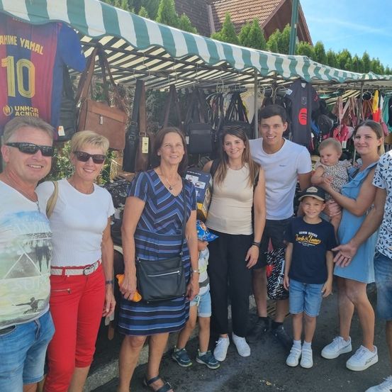 Eine Familie posiert für ein Foto auf einem Outdoor-Markt. Mehrere Gegenstände, darunter Handtaschen und Trikots, hängen an den Ständen.