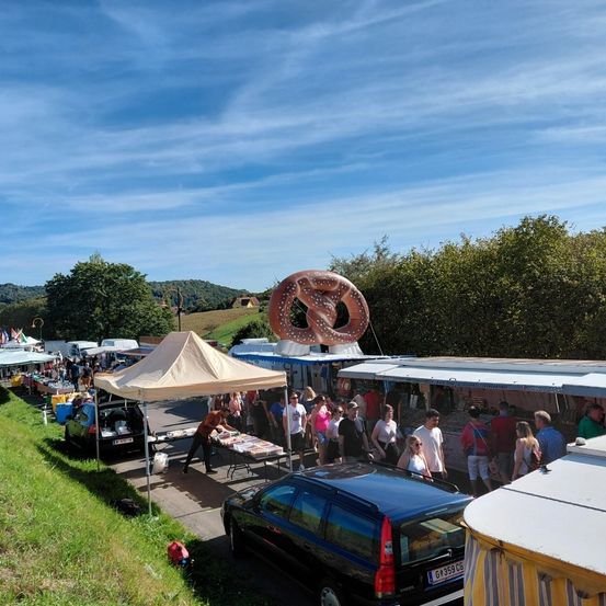 Ein geschäftiger Outdoor-Markt mit mehreren Ständen, einer mit einem riesigen Brezel, und einem klaren blauen Himmel. Leute laufen und shoppen.