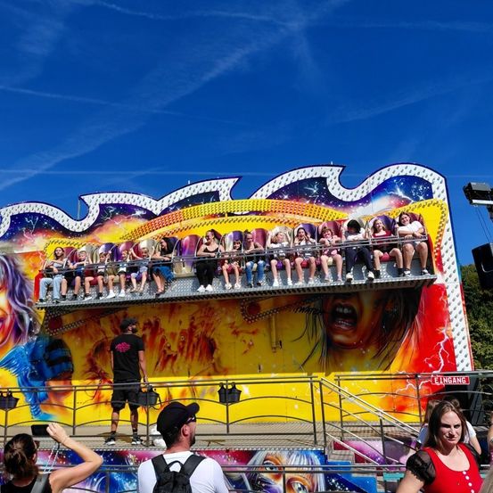 Eine bunte Fahrt mit Menschen in Stühlen, einem Mann in Schwarz neben ihr und einem anderen Mann mit Rucksack, der die Fahrt betrachtet. Die Fahrt hat ein Frauengesicht auf der Seite gemalt.