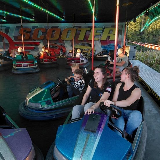 Eine Gruppe von Menschen fährt auf einem Jahrmarkt Bumper Cars. Sie sind unter einem überdachten Bereich mit bunten Lichtern und einem großen Banner.